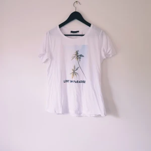 Vit t-shirt med palmtryck från Pine Ridge strl M - Vit t-shirt från Pine Ridge med tryck av palmer och texten 'LOST IN PARADISE' på framsidan. Klassisk rund halsringning och korta ärmar. Skön och lätt i bomull, perfekt för en chill sommarlook.  Helt & fint skick
