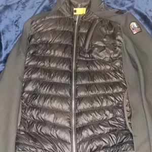 Svart Parajumpers pufferjacka - Snygg svart pufferjacka från Parajumpers med quiltad framsida och stickade ärmar. Jackan har dragkedja, bröstficka med zip och den klassiska gula detaljen vid halsen. Perfekt för kyliga dagar och riktigt stilren look.