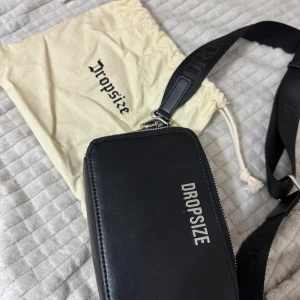 Svart axelväska från Dropsize - Snygg svart axelväska från Dropsize i slätt skinn med silverfärgad logga framtill. Väskan har en bred, justerbar axelrem med broderad text och dragkedja upptill. Kommer med en beige dustbag med logga. Perfekt för att bära det viktigaste med stil.