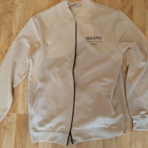 Beige Milano ziptröja  - Snygg beige ziptröja med svart dragkedja och tryck 'MILANO ITALIA' på både bröstet och ryggen. Jackan har två fickor framtill och en stilren, enkel design. Perfekt för dig som gillar clean streetstyle med italienska vibbar från modestaden Milano 