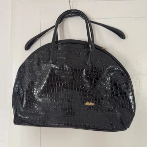 Handväska - Snygg svart handväska från Saba i glansigt lackmaterial med croco-mönster. Väskan har rundad form, två handtag och guldfärgad logga framtill. Baksidan har en praktisk dragkedjeficka. Perfekt för dig som vill sticka ut med en trendig accessoar.
