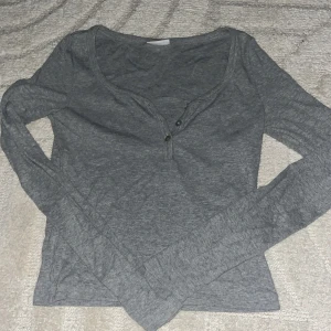 Grå långärmad topp med knappar - Säljer en enkel och stilren grå långärmad topp med två knappar vid halsringningen. Tröjan har en smickrande v-ringning och är gjord i mjuk bomull, perfekt för lager-på-lager eller chill dagar.