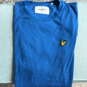 Blå t-shirt från Lyle & Scott - Blå t-shirt från Lyle & Scott med klassisk rund hals och gul broderad logga på bröstet. Tillverkad i mjuk bomull för en skön känsla. Perfekt för dig som gillar stilrena och enkla plagg med en snygg detalj.