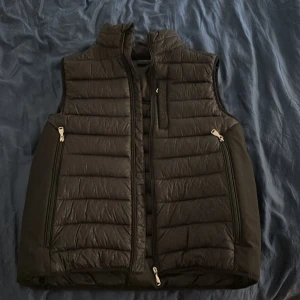 Svart dunväst  - Snygg svart dunväst med quiltad design och hög krage. Västen har tre dragkedjeförsedda fickor framtill och en diskret logga på ryggen. Perfekt att slänga över en hoodie för extra värme. Klassisk och stilren look.