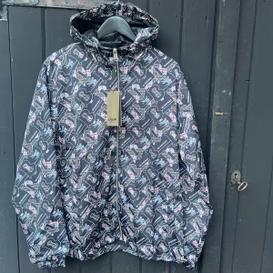 Burberry vindjacka med logomönster - Snygg svart vindjacka från Burberry med heltryckt logomönster i vitt, grått och rött. Jackan har huva med snörning, dragkedja framtill och elastiska muddar. Tillverkad i glansig polyester som ger en modern vibe. Perfekt för dig som vill sticka ut med stil.