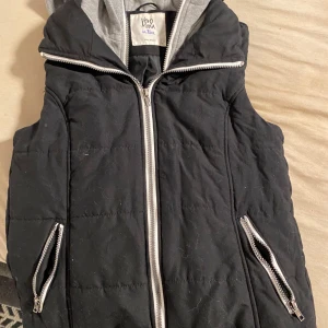 Svart dunväst från Vero Moda - Svart quiltad dunväst från Vero Moda med grå huva och silvriga dragkedjor framtill och vid fickorna. Västen har en sportig look och passar perfekt till lager-på-lager-stil under kyliga dagar.