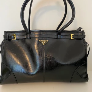Svart handväska från Prada i skinn - Snygg svart handväska från Prada i glansigt skinn med klassisk rektangulär form. Väskan har dubbla handtag, guldfärgade metalldetaljer och Pradas ikoniska trekantiga logga framtill. Insidan är fodrad med mönstrat tyg och har en elegant gulddetalj. Inte fast pris, kan diskuteras 