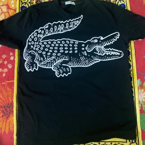 Svart Lacoste t-shirt med krokodiltryck - Cool svart t-shirt från Lacoste med ett stort vitt krokodiltryck på framsidan. Klassisk passform och rund halsringning, perfekt för dig som vill sticka ut med en unik design. Materialet känns mjukt och skönt mot huden.
