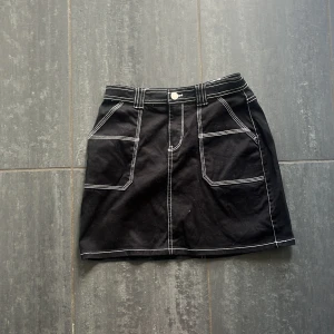 Svart kortkjol med kontrastsömmar Lindex - Svart kortkjol från Lindex med vita kontrastsömmar och stora fickor framtill. Kjolen har bälteshällor och knapp i midjan, vilket ger en cool och stilren look. 