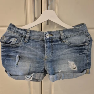 Blå slitna jeansshorts från Lindex - Snygga blå jeansshorts från Lindex med slitna detaljer och upprullade benslut. Klassisk femficksmodell med knapp och dragkedja framtill. Perfekta för varma dagar när du vill ha en avslappnad och trendig look. Det vänstra hålet ska egentligen se ut som de högra bara att de vita trådarna är borta.