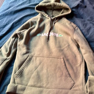 Beige hoodie från Lacoste med huva - Snygg beige hoodie från Lacoste med broderad logga på bröstet och klassisk känguruficka. Tröjan har huva med snörning och är perfekt för en chill och avslappnad stil. Passar dig som gillar streetwear och vill ha något bekvämt och stilrent.