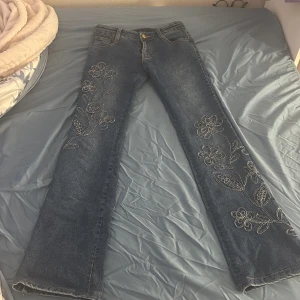Blå jeans med blommig brodyr - Snygga blå jeans med blommig brodyr längs båda benen. Modellen har låg midja och raka ben, perfekt för dig som gillar en chill och trendig look. Jeansen har klassiska fickor och är gjorda i mjukt denimtyg.är 160 ish