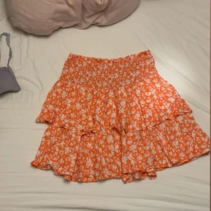 Orange blommig volangkjol Samsa - Superfin kort kjol från Samsa Collection i orange med vitt blommönster. Kjolen har volanger och resår i midjan för en bekväm passform. Perfekt för dig som gillar färg och vill sticka ut lite extra i sommar. Behövs strykas 