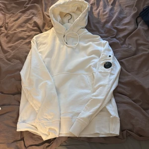 Vit hoodie från C.P. Company - Snygg vit hoodie från C.P. Company magficka och ribbade muddar. Tillverkad i mjuk bomull, perfekt för dig som gillar streetwear och stilrena plagg.                                                         Den har aldrig kommit till någon användning och är 100% äkta.                                            Kom med BUD!                                                                            (Obs säljaren står för frakten) bara att kontakta ifall ni har frågor eller annat!