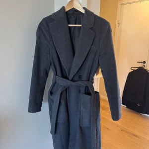 Grå kappa från Tiger of Sweden  - Elegant grå kappa från Tiger of Sweden med klassisk design. Kappan har långa ärmar och ett bälte i midjan för en skräddarsydd look. Den är försedd med knappar framtill och har en stilren krage. (Pris kan diskuteras)