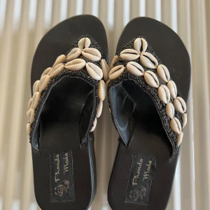 Svarta sandaler med snäckdetaljer - Svarta sandaler med platå från Phanda Moda dekorerade med vita snäckor. Perfekta för en somrig look med en touch av bohemisk stil. Sandalerna har en bekväm sula och en stilren design som passar till många olika outfits.