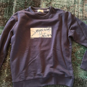Mörkblå tröja från Suspicious Antwerp - Säljer en stilren mörkblå tröja från Suspicious Antwerp med tryck på framsidan. Tröjan har långa ärmar och rund halsringning. Perfekt för en avslappnad look.