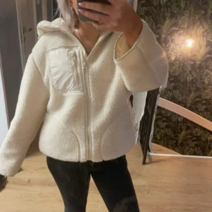 HM Teddyjacka - Mysig vit teddyjacka med huva och dragkedja framtill. Jackan har en praktisk bröstficka med dragkedja och långa ärmar. Perfekt för kyliga dagar när du vill hålla dig varm och bekväm.❤️ stor i storlek 