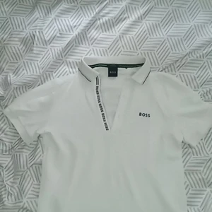 Vit kortärmad t-shirt från Boss - Säljer en stilren vit t-shirt från Boss med korta ärmar och klassisk krage. T-shirten har en diskret logga på bröstet och en snygg detalj med text längs knäppningen. Perfekt för en avslappnad men ändå elegant look.