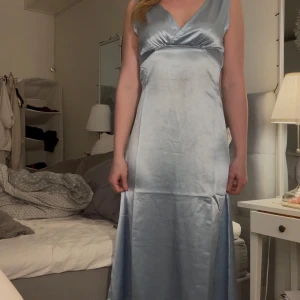 Ljusblå långklänning i satin - Elegant ljusblå långklänning i satin med djup v-ringning och öppen rygg. Klänningen är ärmlös och har en vacker glans som ger en lyxig känsla. Perfekt för speciella tillfällen som exempelvis balen!