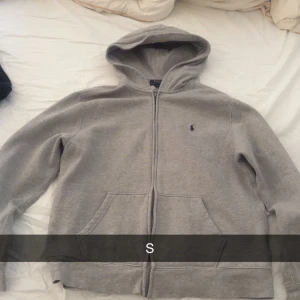 Grå hoodie från Ralph Lauren - Säljer en klassisk grå hoodie från Ralph Lauren med dragkedja och kängurufickor. Den har en liten broderad logga på bröstet och en bekväm huva. Perfekt för en avslappnad stil.