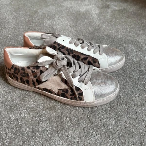 Vår skor - Snygga sneakers med leopardmönster och metallicdetaljer. Skorna har en stjärndetalj på sidan och snörning framtill. De är i en mix av beige, brun och silver med en vit sula.