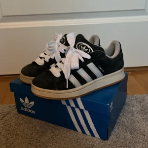 Svarta Adidas Campus sneakers - Snygga svarta Adidas Campus sneakers med vita ränder och breda vita snören. Skorna har en klassisk design med en bekväm sula och är perfekta för en stilren look. De kommer i originalkartong.