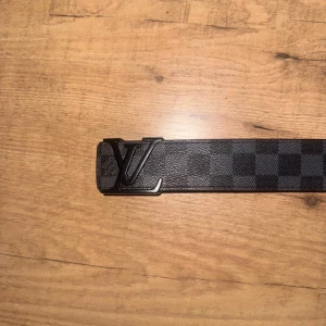 Rutig bälte  - Snyggt bälte från  med ett klassiskt rutigt mönster i svart och grått. Bältet har en stilren svart spänne med -logotypen. Perfekt för att ge en lyxig touch till din outfit.