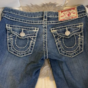  True Religion - Snygga blå jeans från True Religion med kontrasterande vita sömmar och unika bakfickor med knappar. Klassisk femficksdesign och knappgylf. Perfekta för en avslappnad stil. Modellen är raka och förra ägaren har kortat ner längen så det går att förlänga den igen.