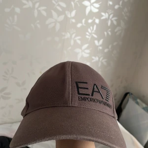 Brun keps från Emporio Armani - Snygg brun keps från Emporio Armani med svart broderad logga framtill. Kepsen är tillverkad i 100% bomull och har en justerbar rem baktill för perfekt passform.