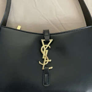 Svart axelväska från YSL - Snygg svart axelväska från YSL med guldfärgad logga framtill. Väskan har en justerbar axelrem och stängs med dragkedja. Perfekt för att ge en lyxig touch till din outfit.