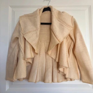 Gul stickad kofta - Säljer en elegant beige stickad kofta med volangdetaljer och öppen design. Koftan har trekvarts ärmar.