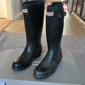 Svarta glittriga stövlar från Hunter - Snygga svarta glittriga stövlar från Hunter. Perfekta för regniga dagar med en stilren design och hög skaft. De har en justerbar rem på sidan för bättre passform.