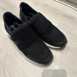 Svarta sneakers från Michael Kors - Snygga svarta sneakers från Michael Kors med elastiska band och nätmaterial. Skorna har en diskret logga på sidan och en glittrig häl. 