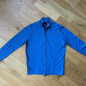 Fullzip merinoulltröja från Oscar Jacobson - Säljer en stilren blå tröja i 100% merinoull från Oscar Jacobson. Tröjan har en praktisk dragkedja framtill och långa ärmar. Perfekt för att hålla sig varm och snygg under kyligare dagar. Tillverkad i Italien. Orginal pris 1500 KR