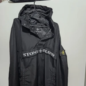 Svart jacka från Stone Island - Snygg svart jacka från Stone Island med huva och tryckknappar framtill. Jackan har en stor logotyp på bröstet och en ikonisk patch på ärmen. Perfekt för en stilren look. Den är jätte skön och priset kan diskuteras vid snabb äffar