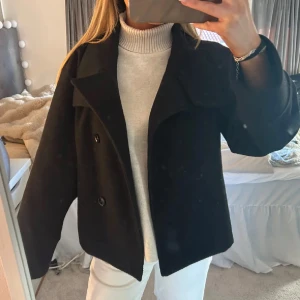 Svart kappa - Snygg svart kappa med dubbelknäppning och bred krage. Perfekt för kyligare dagar och ger en stilren look. Passar bra över en stickad tröja för extra värme.