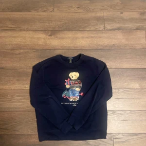 Mörkblå tröja med nallemotiv från Polo Ralph Lauren - Säljer en mörkblå tröja från Polo Ralph Lauren med ett charmigt nallemotiv på framsidan. Tröjan har långa ärmar och en klassisk rund halsringning. Perfekt för en avslappnad stil med en touch av lekfullhet.