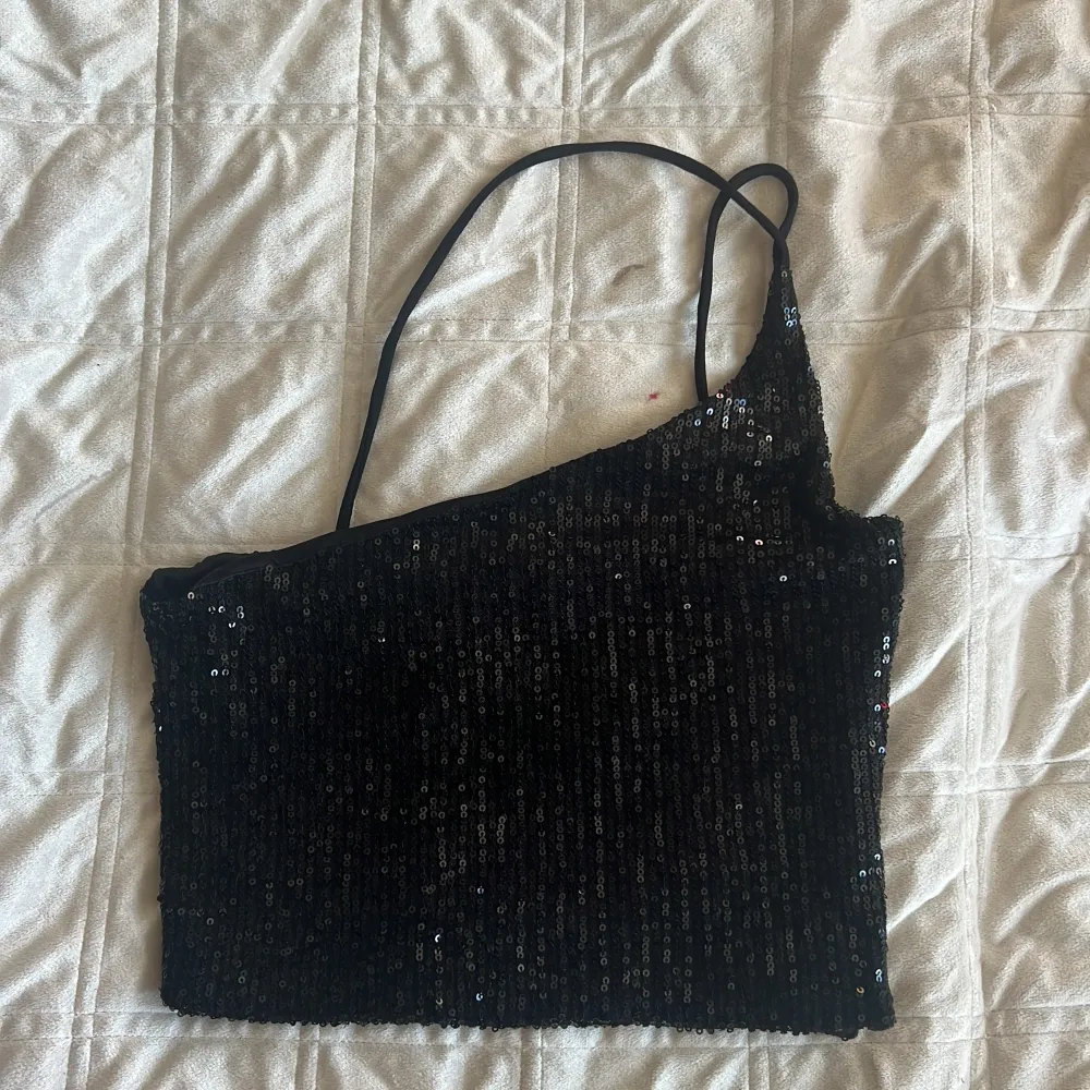 Snygg svart oneshoulder topp med paljetter som ger en glittrig look. Perfekt för festliga tillfällen. Den har en smal axelrem och en asymmetrisk design som ger en modern touch.. Yläosat.