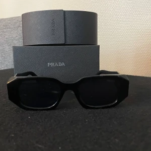 Svarta solglasögon från Prada - Snygga svarta solglasögon från Prada med en rektangulär form. Glasögonen har en elegant och stilren design med märkets logotyp på sidan. Perfekta för att ge en lyxig touch till din outfit.