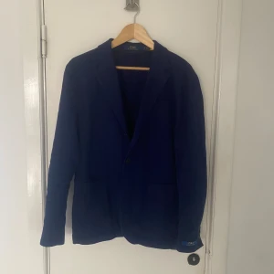 Blå kavaj från Polo Ralph Lauren - Snygg blå kavaj i skön linnemix från Polo Ralph Lauren. Klassisk design med två knappar framtill och fickor. Perfekt för både formella och avslappnade tillfällen. Ett tidlöst plagg som passar i garderoben året runt. Nypris ligger på runt 6000kr. Storlek M och passar storlek 50. Skriv om ni har några övriga frågor🙌