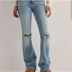 Ljusa jeans med slitningar - Säljer ett par ljusa jeans med slitningar på knäna. De har en bootcut-stil och är perfekta för en avslappnad look. Jeansen har en klassisk femficksdesign och är tillverkade i denim.