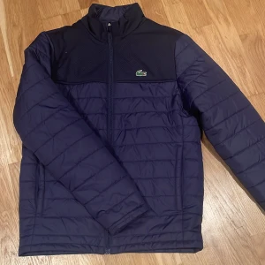 Mörkblå jacka från Lacoste Sport - Säljer en stilren mörkblå jacka från Lacoste Sport. Jackan har en dragkedja framtill och det ikoniska krokodilmärket på bröstet. Perfekt för kyligare dagar med sin quiltade design och långa ärmar.