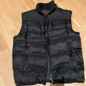 Svart dunväst från Adventure - Säljer en svart dunväst från Adventure med dragkedja och knappar. Västen har två fickor med dragkedja framtill och en hög krage för extra värme. Perfekt för kyliga dagar.