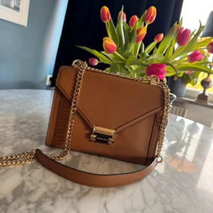 Brun axelväska med kedja - Snygg brun axelväska från Michael Kors med guldfärgad kedja och elegant låsdetalj. Väskan har en stilren design med ett praktiskt innerfack med dragkedja. Perfekt för att bära dina nödvändigheter med stil.