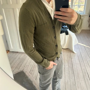 Olivgrön Cardigan från Nudie Jeans - Säljer en stilren olivgrön kofta från Nudie Jeans. Den har en klassisk design med knappar framtill och ribbade kanter. Perfekt för lager-på-lager under kyligare dagar.