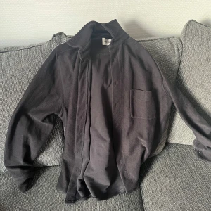 Svart overshirt - Säljer en stilren svart cardigan jacka från East West. Jackan har en enkel design med en öppen front och en bröstficka. Perfekt för lager-på-lager och passar till många olika stilar.