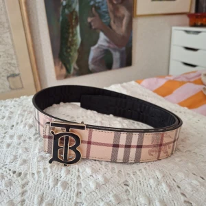 Vändbart burberry bälte - Säljer ett vändbart Burberry-bälte i skinn med klassiskt rutigt mönster på ena sidan i beige, rött och svart samt svart rutigt mönster med präglad riddare på andra sidan. Stort TB-spänne i svart och guld ger en lyxig vibe. Perfekt accessoar för att lyfta din outfit.