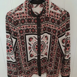 Mönstrad blus från Zara  - Snygg blus från Zara i storlek S med unikt rött, svart och beige mönster. Skjortan har långa ärmar, klassisk krage och knäppning framtill. Materialet känns lätt och flowy, perfekt för dig som gillar statement-plagg med coola detaljer.