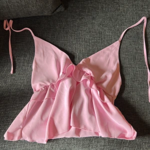 Rosa volang topp med knytband - Supersöt ljusrosa linnetopp med volangdetaljer och smala knytband som går att justera. Toppen har v-ringning och är tillverkad i mjuk polyester, perfekt för varma dagar. Passar dig som gillar en feminin och luftig stil.